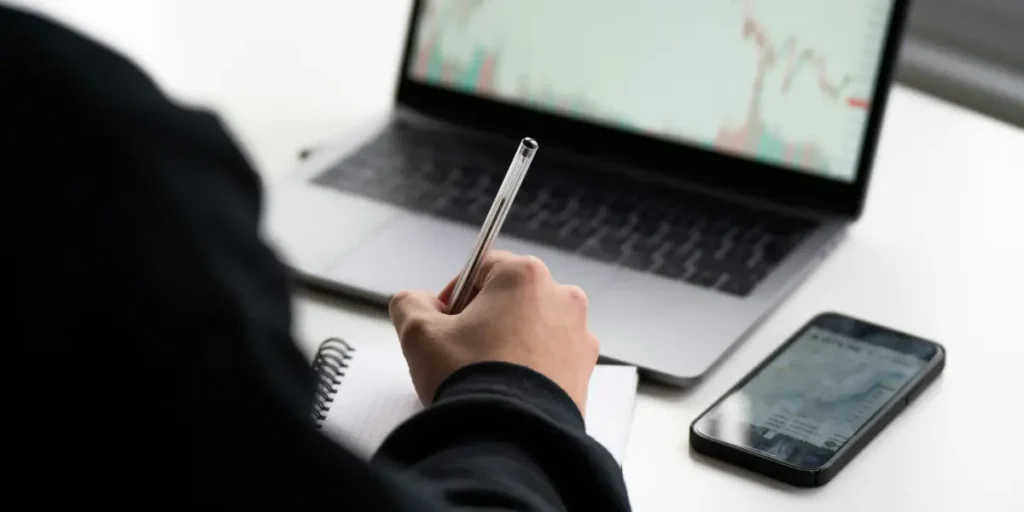 Person Taking Notes While Reviewing Crypto Market Charts On A Laptop With A Smartphone Nearby, Researching Denver Digital Marketing Strategies And Seo Insights.