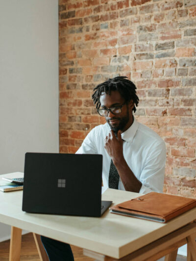 Man sitting in open office area working on his laptop. Wood desk, brick background.