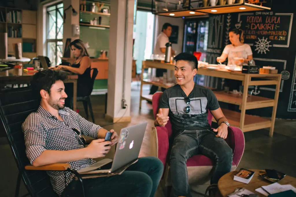 Two men smiling and talking in a coffee shop while one uses a laptop