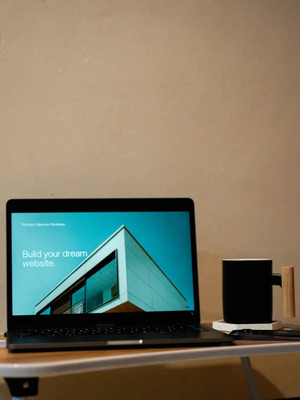 Laptop screen displaying “Build your dream website” with a modern building image beside a coffee mug on a desk