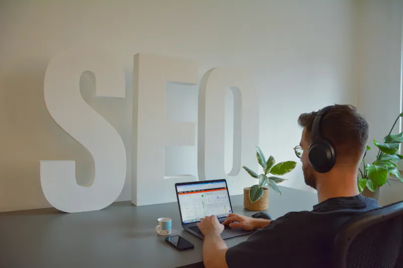 Man wearing headphones working on SEO analytics at a laptop with large SEO letters behind him